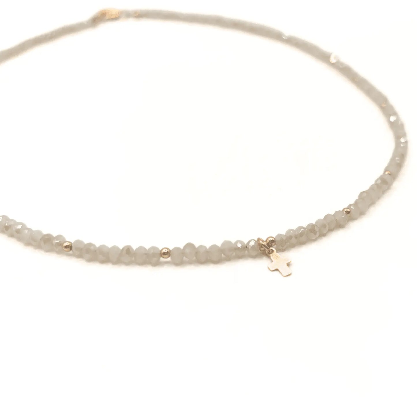 The Gold Filled Little Luxe Cross Necklace On Winter White Crystal Beads features small translucent gray beads, gold accents, and a tiny gold filled cross charm at the center. The waterproof necklace is shown on a white background.