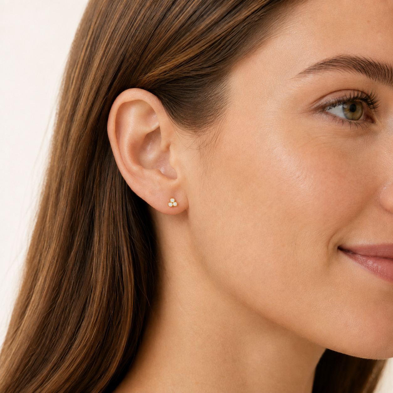 A close-up of a woman's ear wearing a small gold stud earring with three tiny clear stones, set against a clean white background.