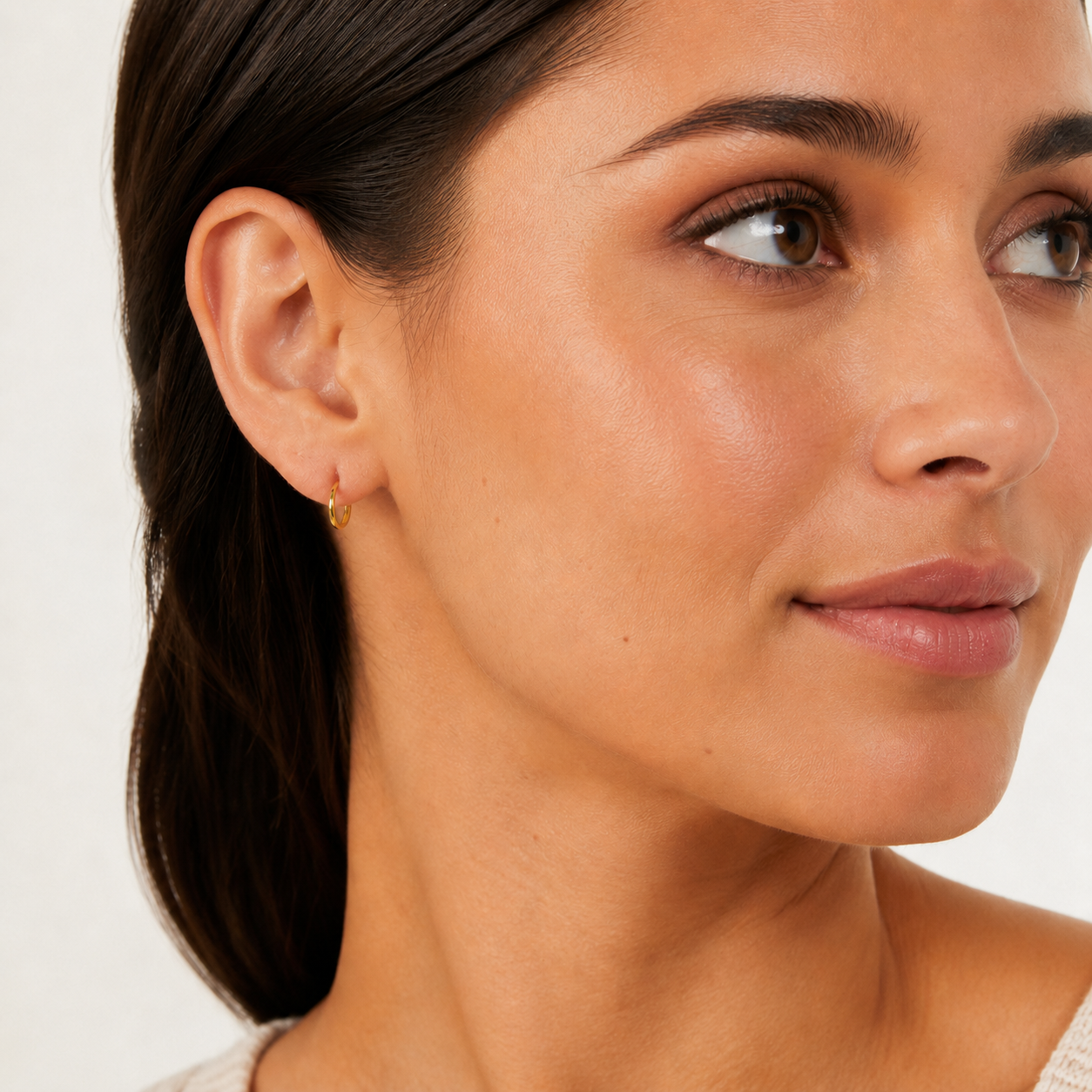 A close-up of a woman's ear wearing a delicate gold huggie hoop earring, shown against a clean white background.
