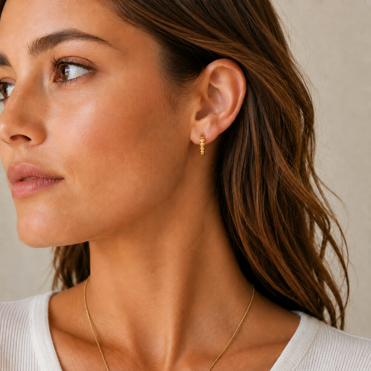 A close-up of a woman wearing a delicate gold huggie hoop earring, paired with a fine gold chain necklace and a white ribbed top, set against a soft beige background.