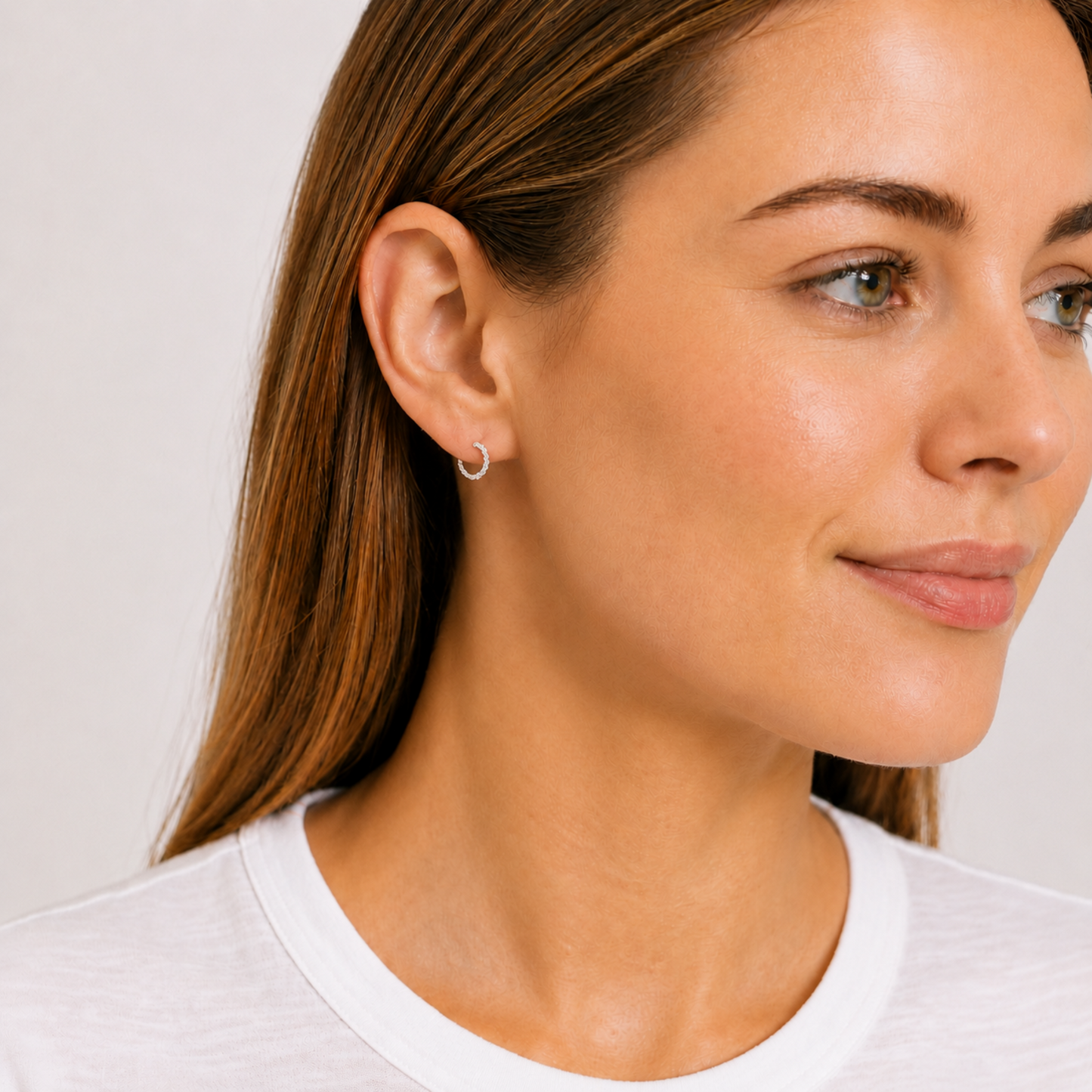 A close-up of a woman wearing a delicate silver hoop earring, shown from the side against a light gray background.