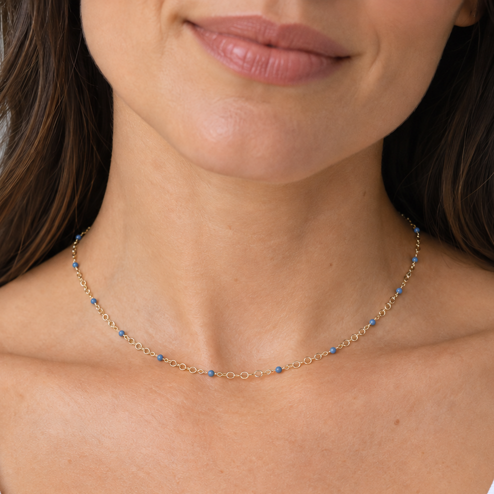 A close-up of a woman wearing a delicate gold chain necklace with small blue bead accents, paired with a white ribbed tank top.