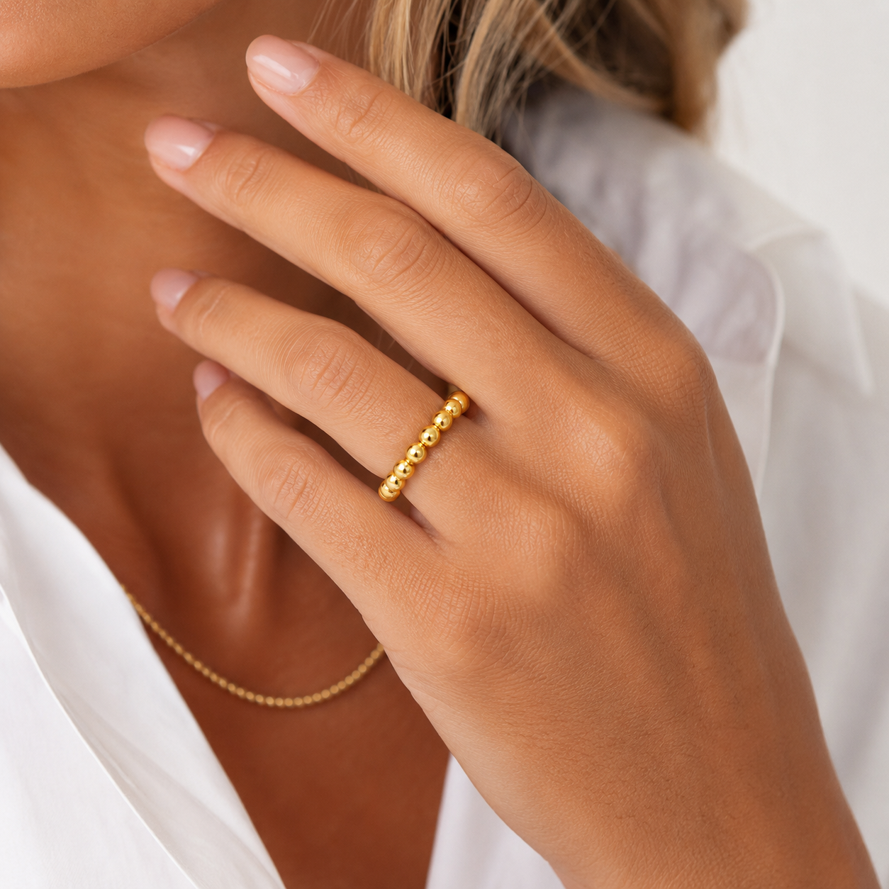 A close-up lifestyle shot of a woman's hand wearing a delicate erin gray gold beaded ring, with a matching gold chain necklace partially visible against a white shirt – erin-gray