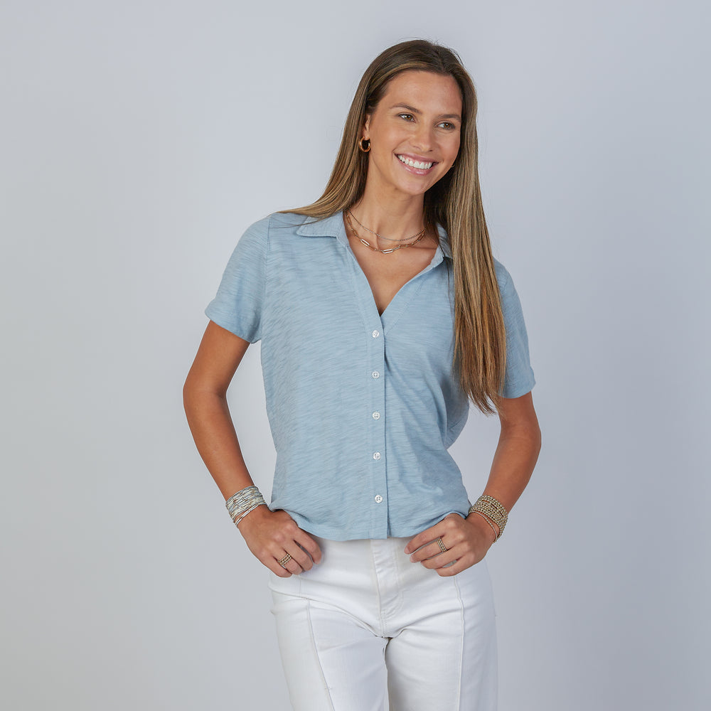 Woman wearing an erin gray design dusty blue Polo short-sleeve button-up shirt and white pants against a plain background