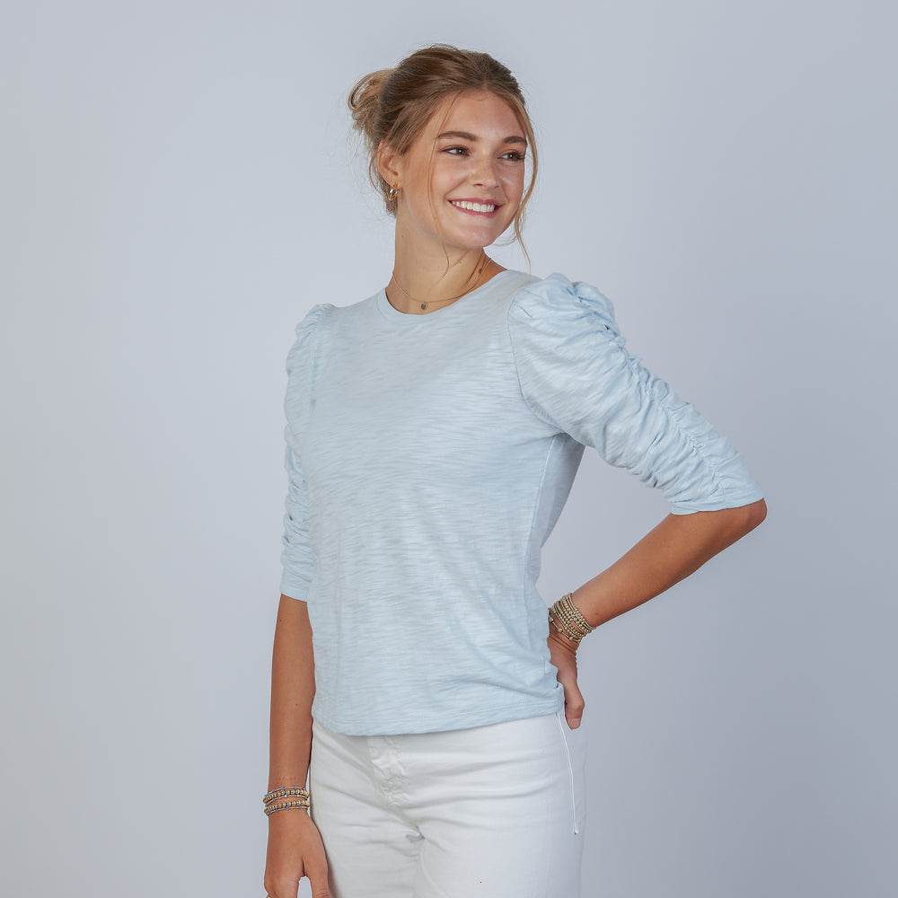 Woman wearing an erin gray design sophie sky blue shirt and white jeans against a gray background